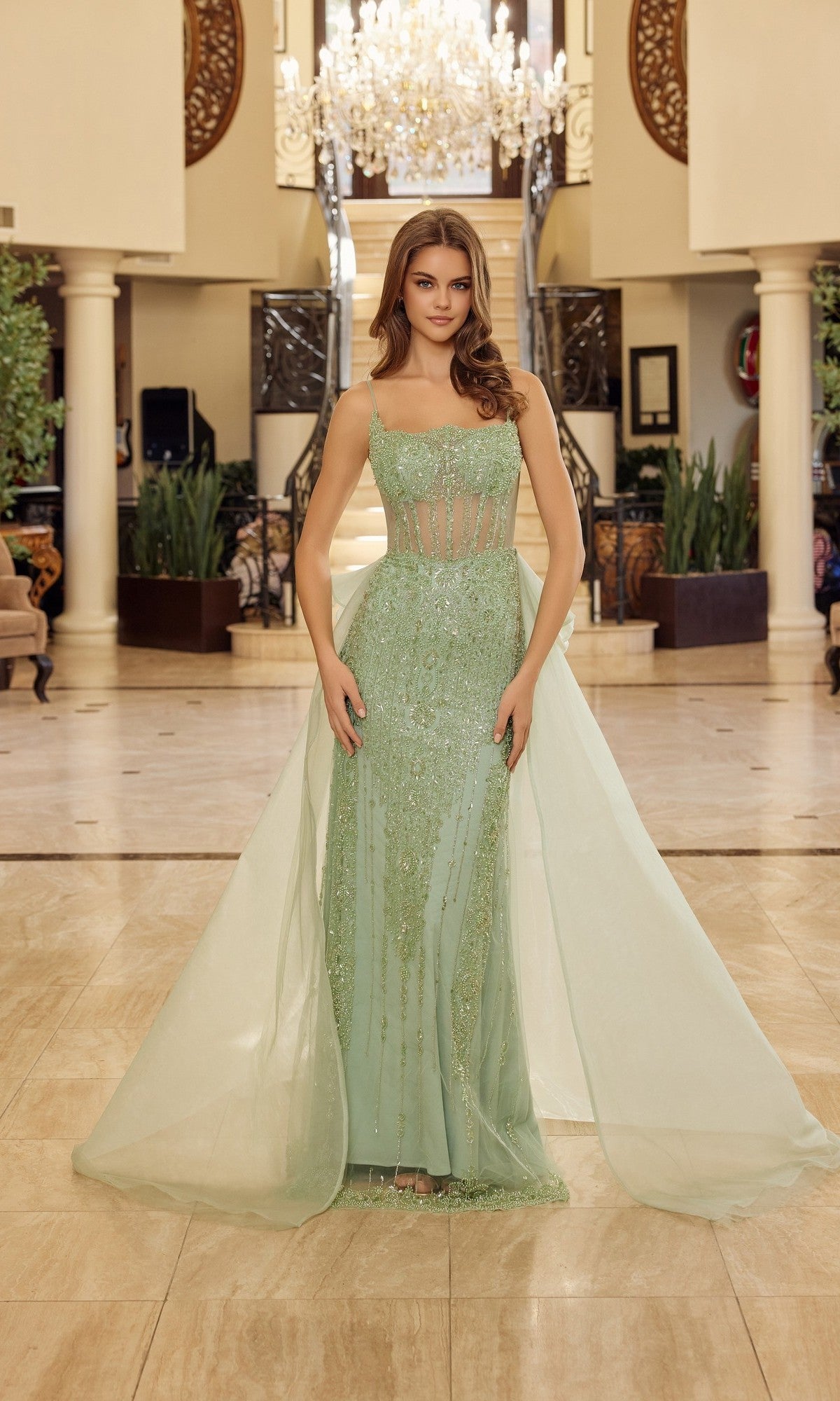 A woman wears the Nox Anabel N1763 long prom dress in sage green, featuring beaded accents, sheer panels, and a sheath silhouette, standing in an elegant foyer with a chandelier and grand staircase.